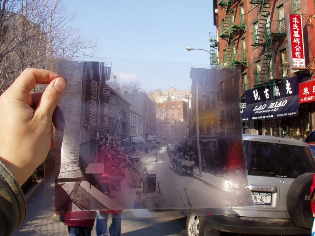 someone standing in a street in nyc, holding up a transparent photograph of the same street at a different time