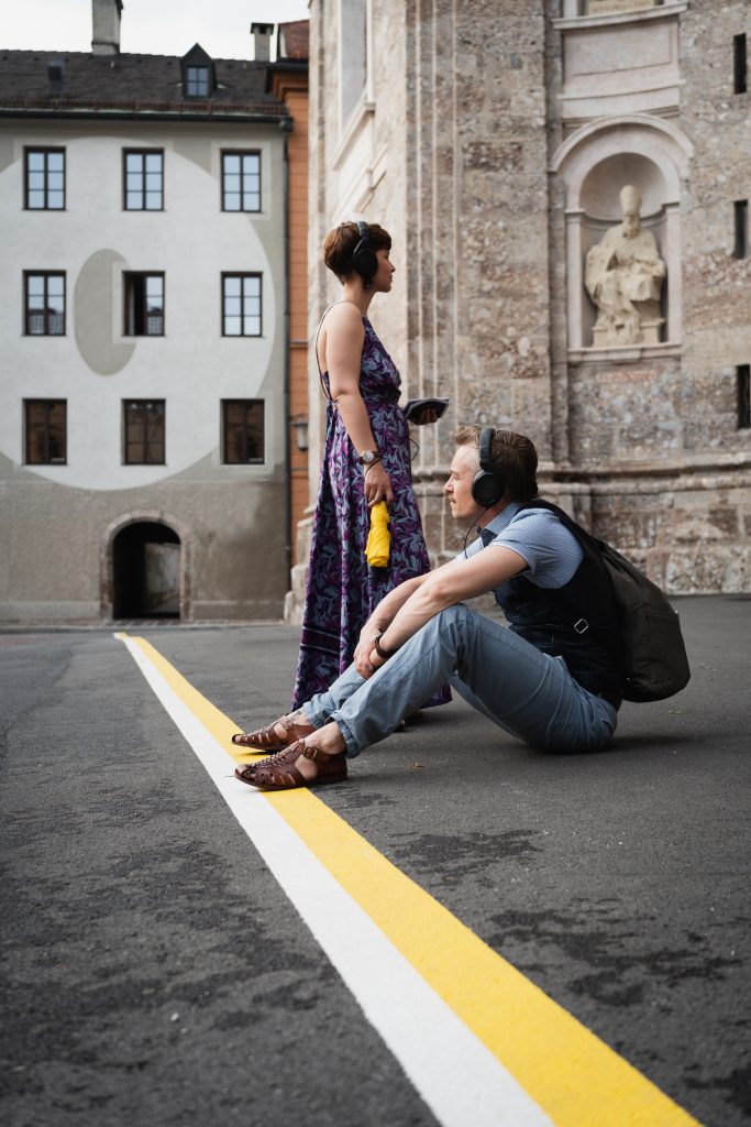 two people with headphones in front of a church. one is sitting on the floor looking away from it, the other standing looking in