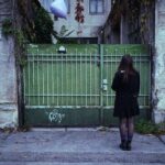 a girl stands in front of a green gate, in her hand a purple heart balloon