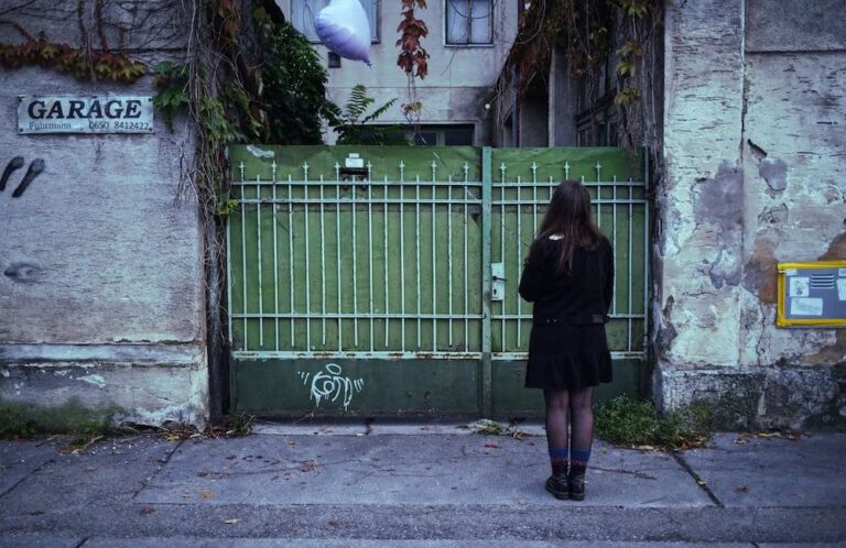 a girl stands in front of a green gate, in her hand a purple heart balloon